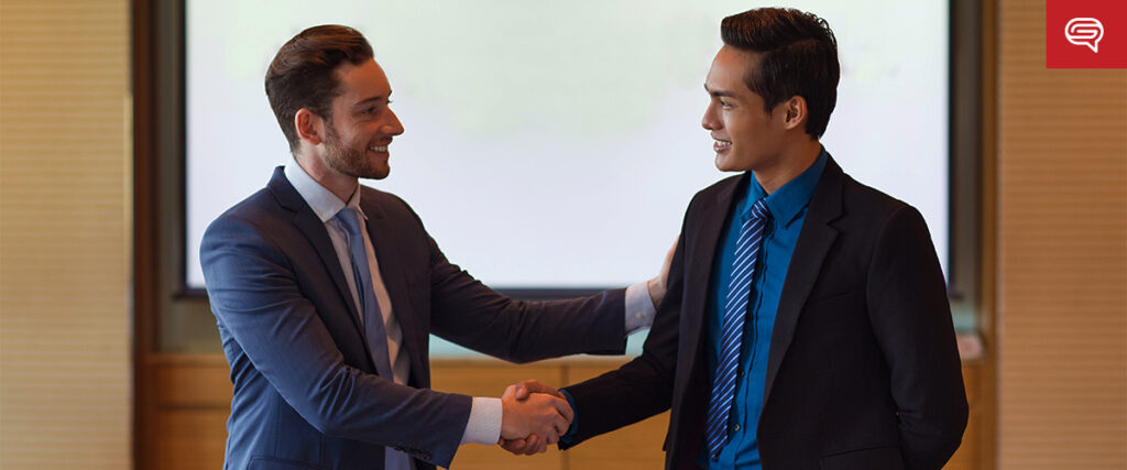 Two men in formal attire are shaking hands and smiling at each other in a professional setting, likely an office or conference room with a large presentation slide in the background. The man on the left is wearing a blue suit, while the man on the right is dressed in a dark suit.