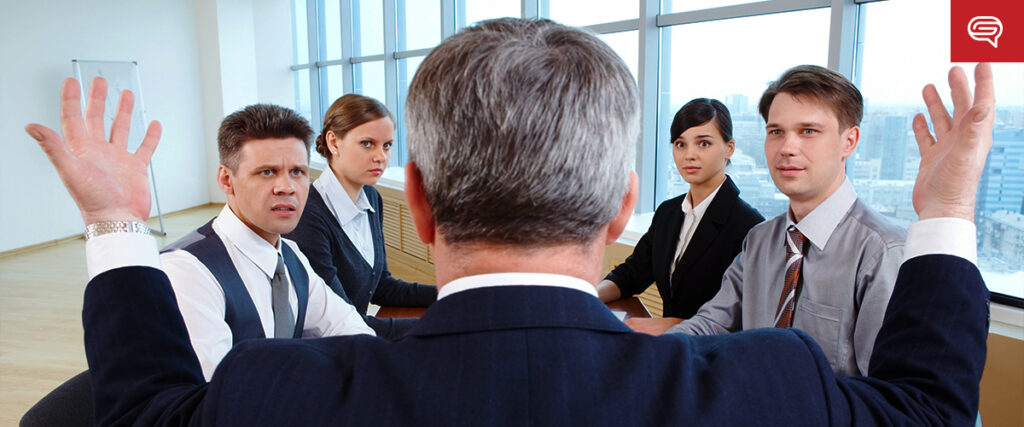 A person in a suit stands with arms raised, facing a table of four seated individuals in a meeting room. The seated individuals, two men and two women, look surprised or confused as they view the pitch deck. The room has large windows, offering a view of the cityscape.