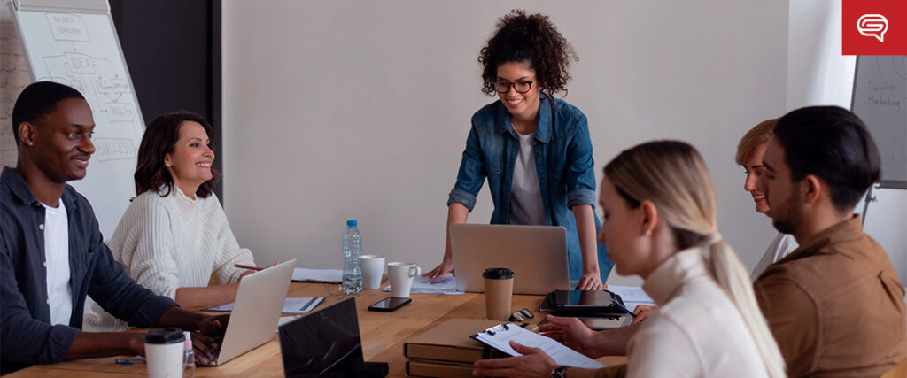 A diverse group of six people are gathered around a large wooden table in a meeting room. Several laptops, papers, and coffee cups are on the table. A woman with curly hair stands while presenting a pitch deck, while the others sit, smiling and engaged. A whiteboard is in the background.