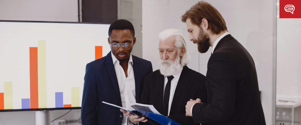 Three men in business attire are gathered, examining documents together. Behind them, a screen displays a colorful bar graph from their pitch deck. The man on the left is wearing glasses and a navy suit, the man in the middle has white hair and a beard, and the man on the right has a beard.