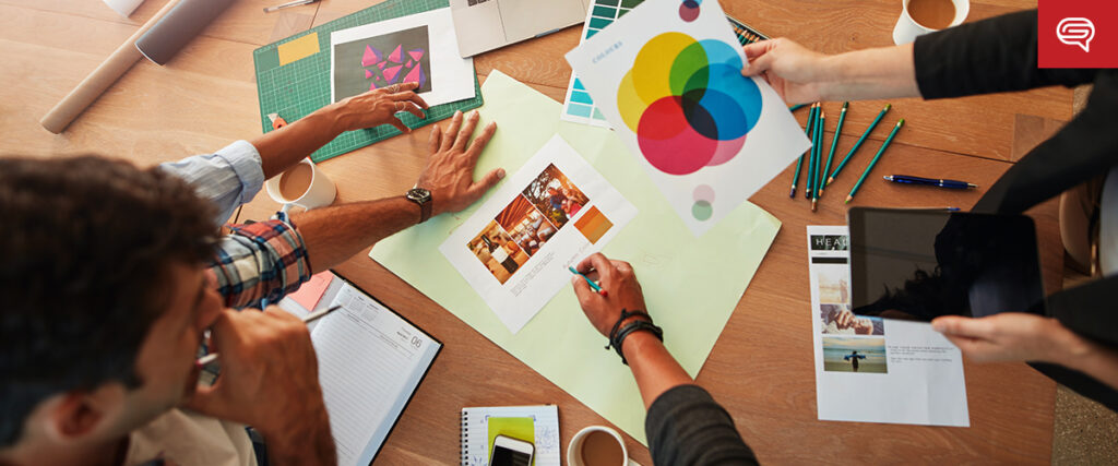 A group of people collaborating on a project at a wooden table. One person is holding a color wheel chart, another is working on a design layout for a PowerPoint presentation, and various tools like pencils, a tablet, and a cutting mat are scattered around. Cups of coffee are also visible.