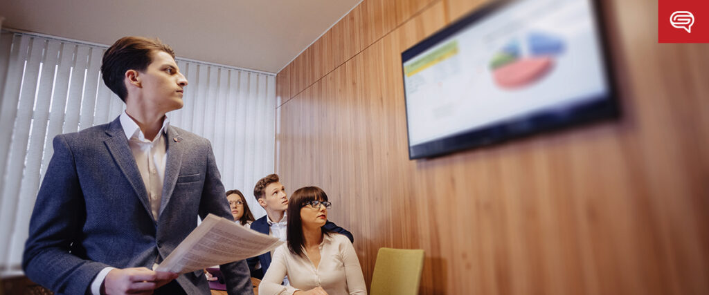 A man in a grey blazer holds papers while presenting data displayed on a slide to seated colleagues in a meeting room. The audience members appear attentive, and the screen shows a pie chart among other charts. The room has wooden walls and window blinds.