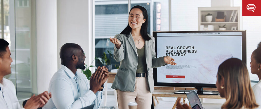 A woman stands in front of a group of colleagues, presenting a slide template that reads "Real Growth, Real Business Strategy" on a large monitor. The group, sitting around a table in the modern office with natural light and shelves in the background, listens and takes notes.