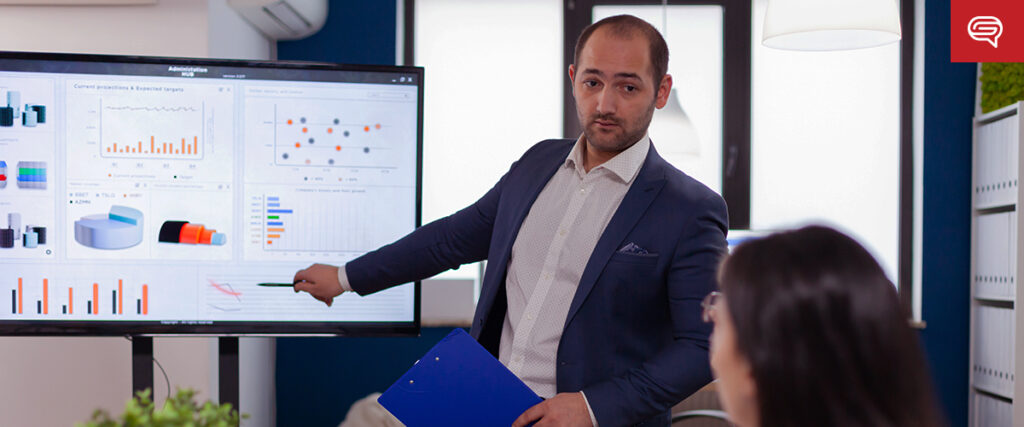 A man in a suit is giving a presentation in an office, pointing to various charts and graphs displayed on a screen. He is holding a blue folder in his other hand. Another person, who is blurred, is seated and looking at the slide.