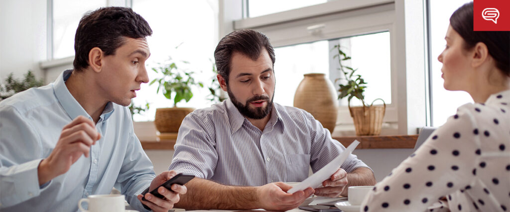 Three people are having a discussion at a table, with documents and a smartphone in hand. Two men are facing a woman. One man is holding papers, while the other gestures about the PowerPoint slide template, and the woman listens attentively. Potted plants are visible in the background.