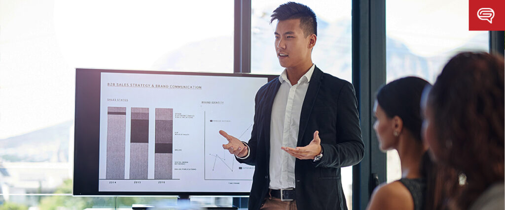 A man is giving a business presentation in a modern office. He stands in front of a screen displaying slides related to sales strategy and brand communication. Two colleagues sit at a table, listening attentively.