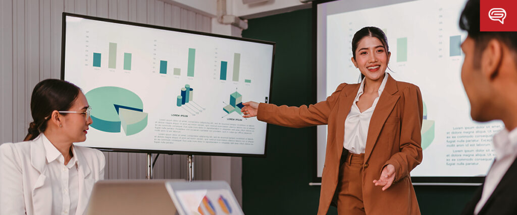 A woman in a brown suit is presenting a PowerPoint business report to colleagues in a meeting room. Two large screens display charts and graphs. Two colleagues, sitting at a table with laptops, are watching and listening attentively.