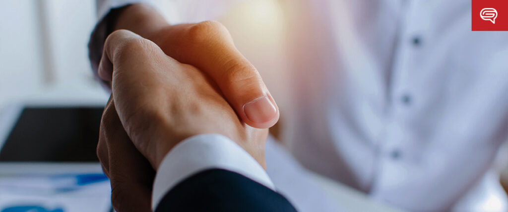 Close-up of two people shaking hands, one in a dark suit and the other in a white shirt. A tablet and some documents on a desk suggest they're discussing a pitch deck. The scene is well-lit, with warm light highlighting the handshake.