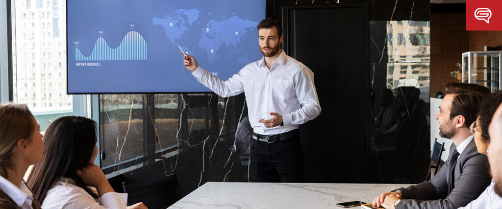 A man in a white shirt stands at the front of a meeting room, pointing to a large screen displaying PowerPoint charts and a world map. Four colleagues sit at a table, listening attentively. The room has a modern design with marble surfaces and large windows.