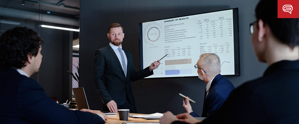 A man in a suit stands next to a screen presenting a PowerPoint slide with charts and data to three colleagues seated at a table. One colleague holds a pen while others listen attentively. A laptop, papers, and coffee cups are on the table in the modern conference room.