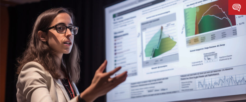 A woman wearing glasses and a light-colored blazer is giving a presentation in front of a large screen displaying complex graphs and charts from her pitch deck. She gestures with her hand towards the screen, emphasizing the data points shown. The background is dimly lit.