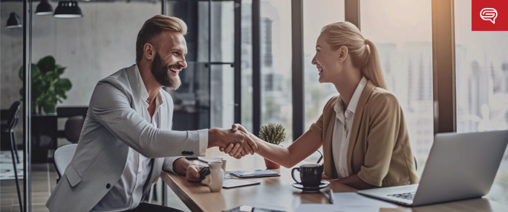 A man and woman sit at a table in a modern office, smiling and shaking hands. There are laptops, coffee cups, and documents on the table. Large windows in the background reveal a cityscape. A pitch deck can be seen on one of the screens as they finalize their discussion.