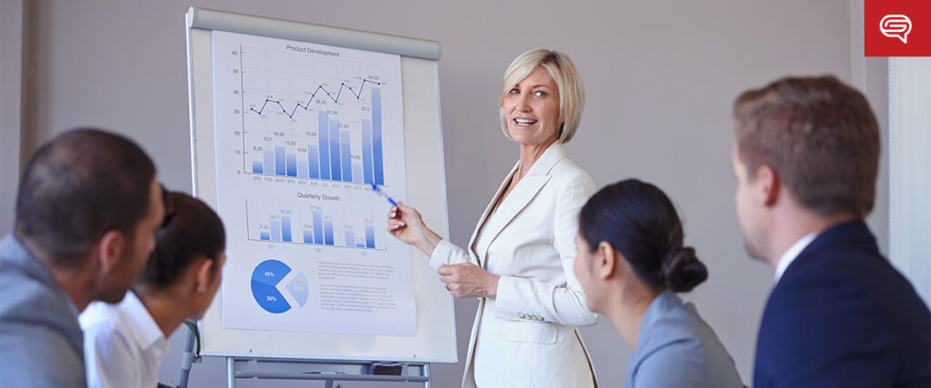 A woman in a white suit gives a presentation to a group of people sitting at a table. She is pointing at a graph on a flip chart displaying project development and quarterly growth data, using her pitch deck. The attendees are attentively listening.