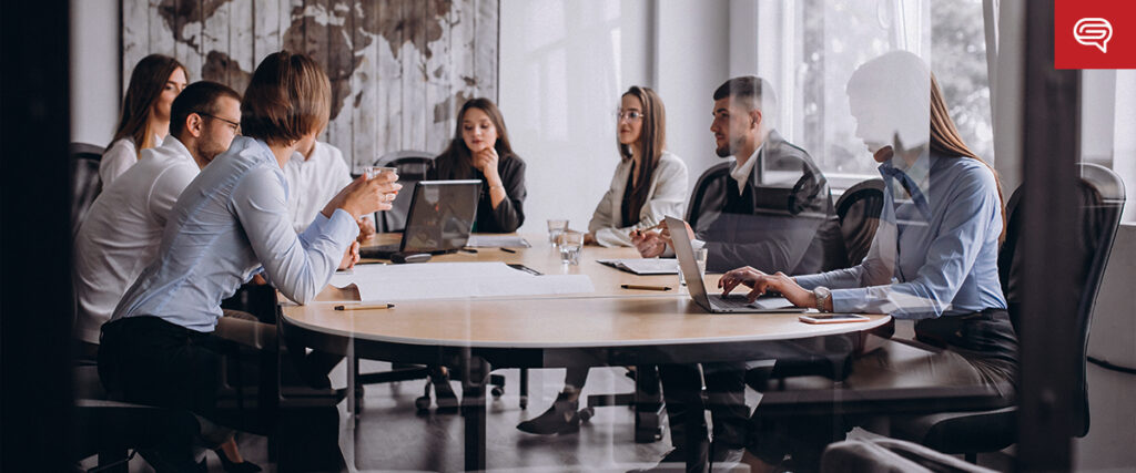 A group of people sit around a long conference table in a modern office, engaging in a meeting. Most are using laptops or taking notes, possibly discussing a slide template. Large windows and minimalistic decor surround them. A map on the wall is partially visible in the background.