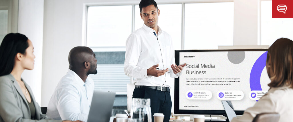 A man stands near a screen giving a presentation on "Social Media Business" to three colleagues seated at a table. The slide template displayed discusses key points on the subject. The background shows a bright office space with large windows.