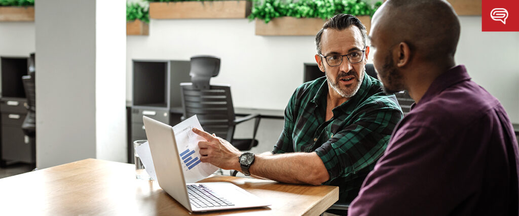 Two men sit at a table in an office setting, discussing documents. One man, wearing glasses and a green plaid shirt, points to a graph on a paper, while the other listens attentively. An open laptop with a PowerPoint slide and glasses of water are on the table.