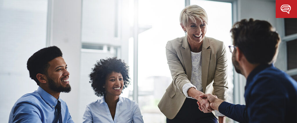 A group of four professionals in a modern office. A smiling woman in a beige blazer is standing and shaking hands with a seated man in glasses, while two other people, a man and a woman, are seated nearby, watching and smiling. They're preparing for an upcoming PowerPoint presentation.