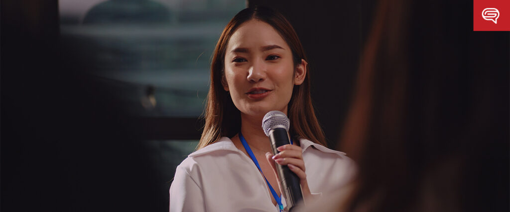 A woman with long hair stands speaking into a microphone, presenting a PowerPoint slide. She is wearing a white shirt and a name tag on a blue lanyard. The background is blurred, focusing attention on her.