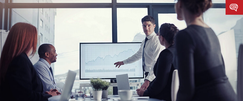 A man in a white shirt and tie points to a line graph on a screen during a business meeting. Four colleagues sit around a conference table listening. The room has large windows, and the atmosphere appears professional and focused as they discuss their pitch deck.