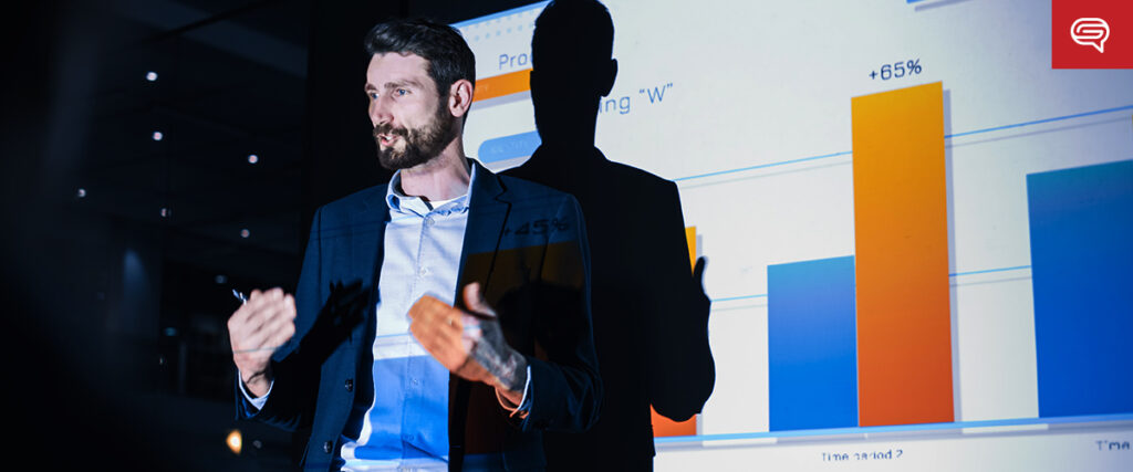 A man in a suit gestures while presenting in front of a projection screen displaying bar graphs with percentage figures and text. The background is dimly lit, emphasizing the bright slide template on the screen. A logo is visible in the top right corner.