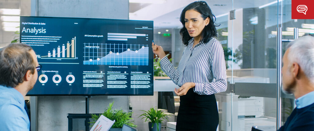 A woman is presenting to two men in an office. She points at a PowerPoint slide displaying various charts, graphs, and the word "Analysis" related to digital technology sales data. The setting is a modern office with glass walls and a bright, professional atmosphere.