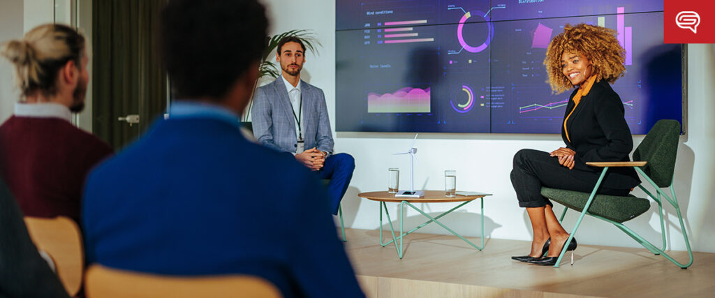 Two presenters are seated in front of a large screen displaying various charts and graphs during a meeting. A woman with curly hair is speaking while a man in a suit listens. Three people are seated in the audience, viewing the slide template presentation.