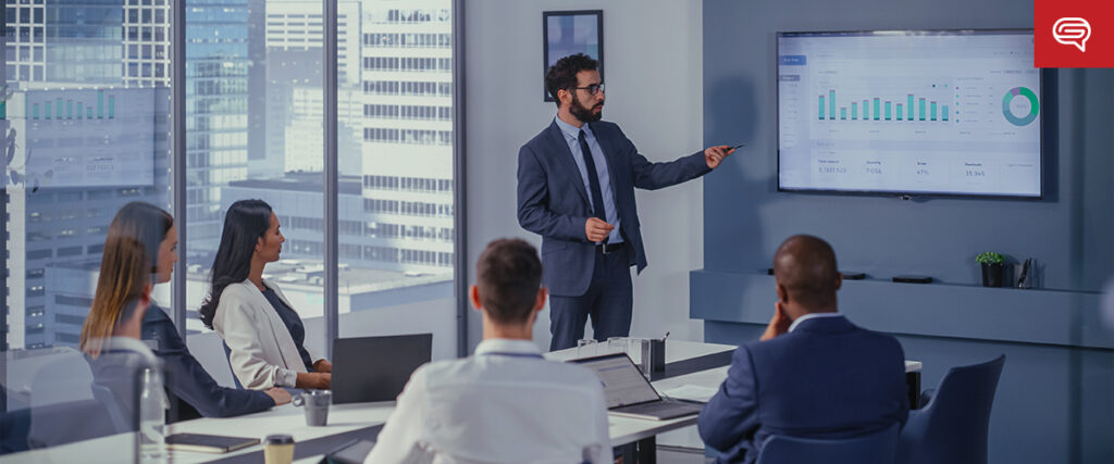 A man in a suit stands and points to a large screen displaying graphs and data in a modern conference room with large windows. Four seated colleagues, two men and two women, listen attentively, taking notes and using laptops. The presentation includes an impressive pitch deck as the urban skyline is visible outside.