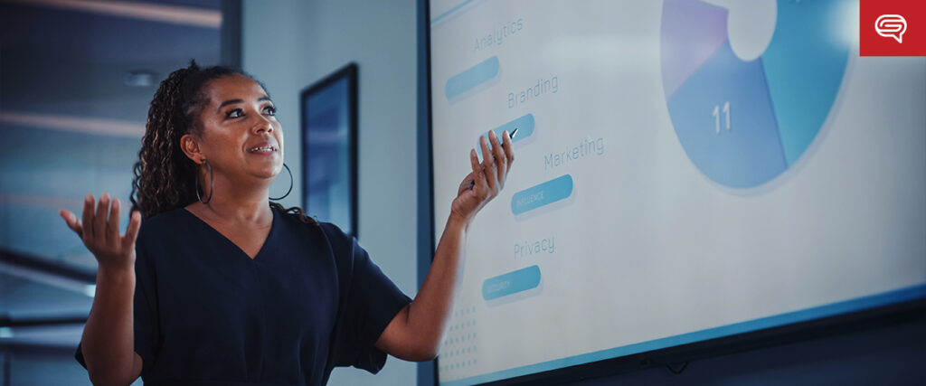 A woman stands in front of a projection screen, gesturing with both hands as she gives a presentation. The screen displays a colorful pie chart and a list of topics like "Analysis," "Branding," "Marketing," "Values," and "Privacy." A logo is in the top-right corner of the PowerPoint slide template.