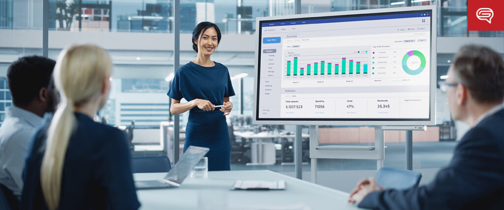 A woman in formal attire presents a PowerPoint pitch deck on a large screen to a group of seated colleagues in a modern office setting. The screen displays various charts and graphs, including bar and pie charts. The office has glass walls and an open, professional environment.