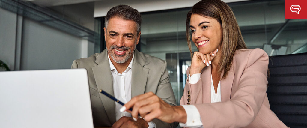 A man in a beige suit and a woman in a pink blazer sit at a desk, smiling and looking at a laptop screen. The woman is pointing at the screen with a pen, possibly discussing a presentation template or collaborating on slides. The background shows a modern office setting.