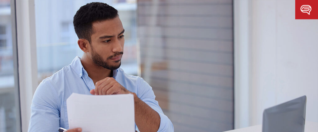 A man with short hair and a beard, wearing a light blue shirt, is sitting at a desk in a bright office. He is focused on examining a pitch deck in his hands while looking at a computer monitor in front of him.