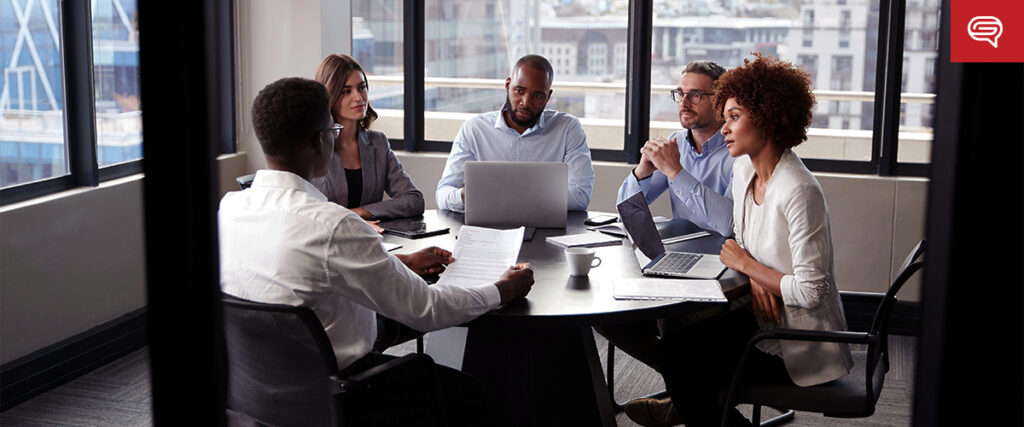 A group of five people are seated around a table in a modern office. They are engaged in a discussion, with laptops, documents, and coffee cups in front of them. Large windows in the background let in ample natural light as they review their pitch deck.