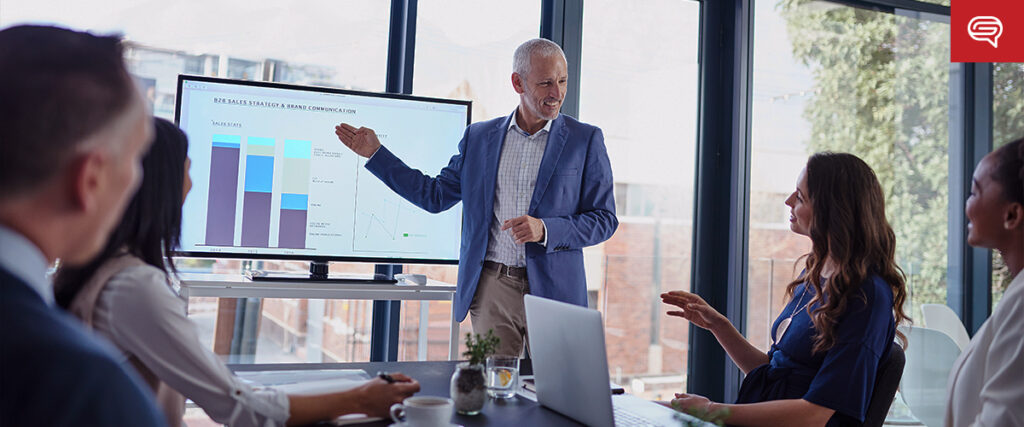 A man in a suit stands in front of a large screen displaying a PowerPoint bar graph and other data, presenting to a group of four seated colleagues in a modern conference room with large windows. The colleagues are listening attentively and have laptops and notebooks.