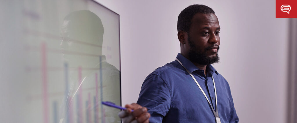 A man in a blue shirt and lanyard stands next to a glass presentation board, holding a pen. He appears to be explaining data or information displayed on the board, similar to a PowerPoint presentation. The setting is likely a professional or educational environment.