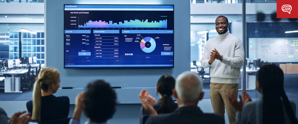 A man stands in front of a large screen displaying data charts and graphs as he presents his pitch deck to an audience in a modern office setting. The attendees are seated and appear engaged, with some applauding. The office space is brightly lit with large windows.