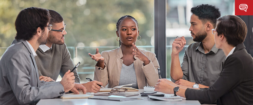 A diverse group of five people sit around a table in a professional setting, engaged in an active discussion. The person in the center is speaking and gesturing with their hands, while the others listen attentively. Various documents and electronic devices, including a pitch deck slide on a tablet, are on the table.