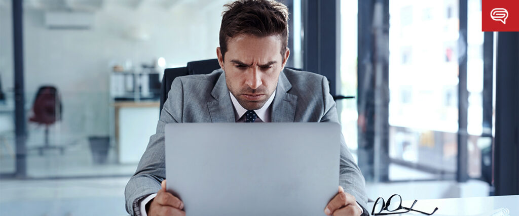 A man in a suit sits at a desk in a modern office, looking intently at his laptop screen. The environment behind him appears professional with large windows and office furnishings. His glasses rest on the desk beside him as he prepares a presentation.