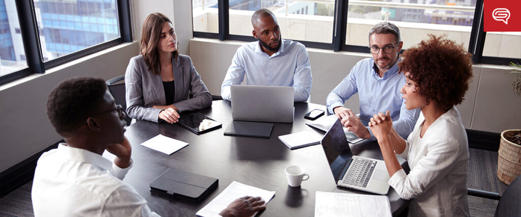 Five people are seated around a conference table in an office, engaged in a meeting. Laptops, tablets, papers, and a cup of coffee are on the table. A presentation is projected on the wall using a slide template. Large windows provide natural light, and a potted plant is in the corner.