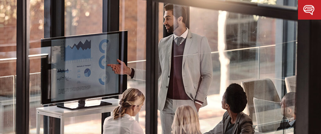 A man in a gray suit is giving a presentation using a slide template on a screen to three colleagues seated around a table in a modern office with large windows. The screen displays graphs and charts. Sunlight is streaming in, creating a bright and focused meeting environment.