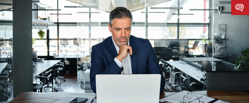 A man in a dark suit sits at a desk in a modern office, looking intently at a laptop. He has a thoughtful expression, resting his chin on his hand, perhaps preparing a presentation. The background shows an open office space with large windows and several desks.