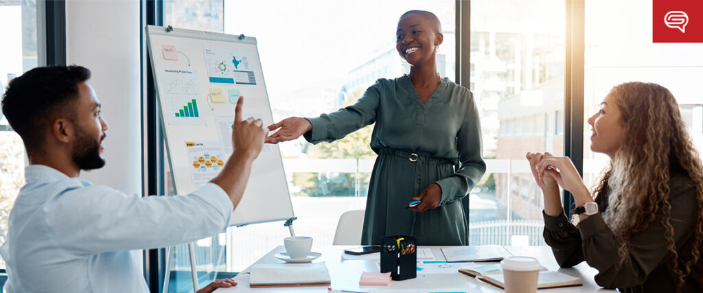 A woman stands next to a flip chart with graphs and charts, smiling and shaking hands with a seated man. Another woman is seated at the table, listening attentively to the presentation. They are in a modern office with large windows.