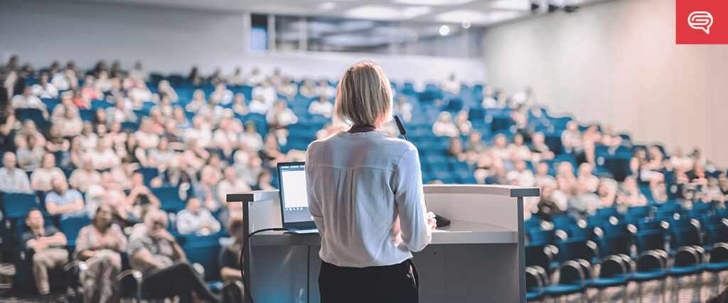 A person with short hair stands at a podium in a large auditorium, delivering a presentation to a seated audience. A laptop with PowerPoint open is on the podium, and the room is filled with rows of blue chairs. The audience is blurred, emphasizing the speaker in focus.