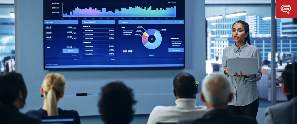 A woman stands in front of a large screen presenting data visuals, including bar graphs and pie charts, to a seated audience in a modern conference room. She holds a laptop, guiding the attendees through the presentation slide by slide as they focus intently on the pitch deck.