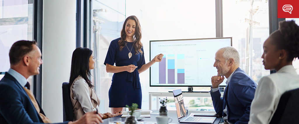 A woman stands next to a large screen displaying slides from a pitch deck as she presents to four colleagues seated around a conference table in a modern office setting. The mood is professional, and all participants appear engaged in the discussion.