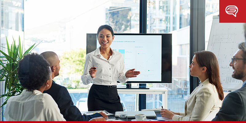 A woman in business attire stands in front of a screen, leading a presentation to four colleagues seated around a table in a modern office. The PowerPoint screen displays charts and graphs, and a flipchart is visible to the right. Large windows show an urban background.