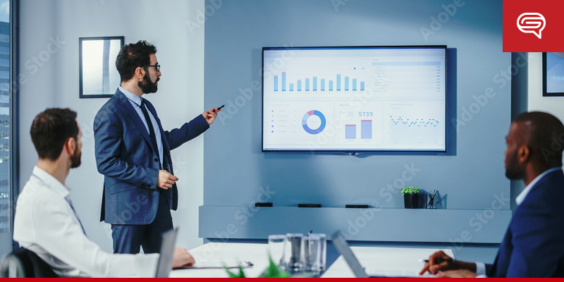 A man in a suit presents data charts on a large screen to three colleagues seated at a conference table. The room has a modern design and is well-lit, featuring a window and plants in the background. The slide template used adds clarity to the presentation.