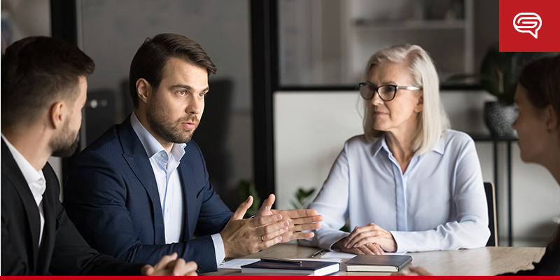 A group of four professionals engaged in a discussion in a modern office setting. A man in a blue suit is speaking, gesturing with his hands, while an older woman with white hair and glasses listens attentively. The atmosphere appears serious and focused.