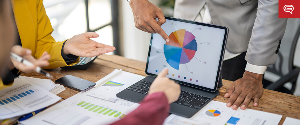 Three people are gathered around a tablet displaying a colorful pie chart. One person is pointing to the slide on the tablet. Documents, charts, and a smartphone are spread out on the wooden table. The setting appears to be a business meeting or presentation.