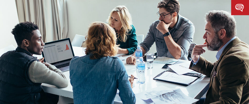 A group of five people engaged in a discussion around a table. One person is working on a laptop, while others have documents and notepads. Water bottles are on the table, and a whiteboard with a red logo is partially visible in the background, possibly preparing for their presentation.