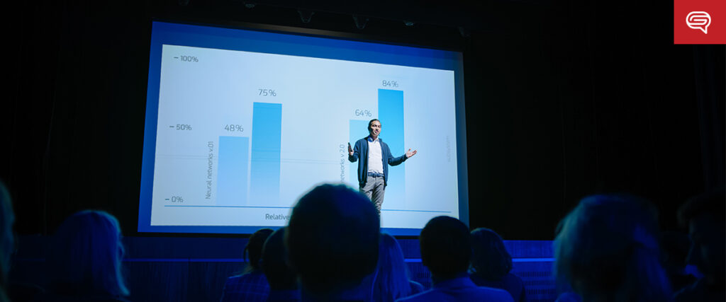 A speaker stands on a stage in front of a large screen displaying a PowerPoint slide with a bar graph. The graph shows three bars with percentages: 75%, 64%, and 84%. Audience members in the foreground listen attentively. The setting appears to be a conference or seminar.